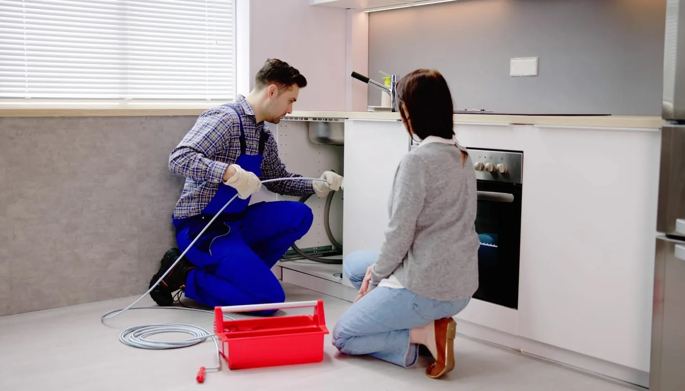 A plumber using a drain snake on a kitchen drain while a homeowner watches